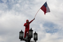 <p>Una persona sobre una farola, vestida como los protagonistas de la Casa de Papel, alza una bandera durante las protestas en Santiago de Chile en 2019.</p>